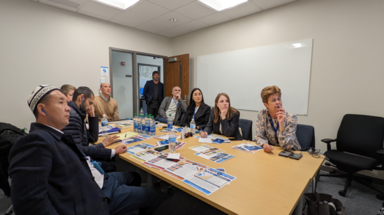 A group sits around a table listening to a video or online presentation.