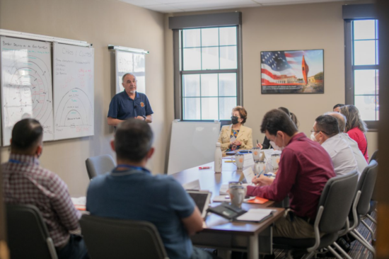 Man standing at board presenting while group of people sit listening.