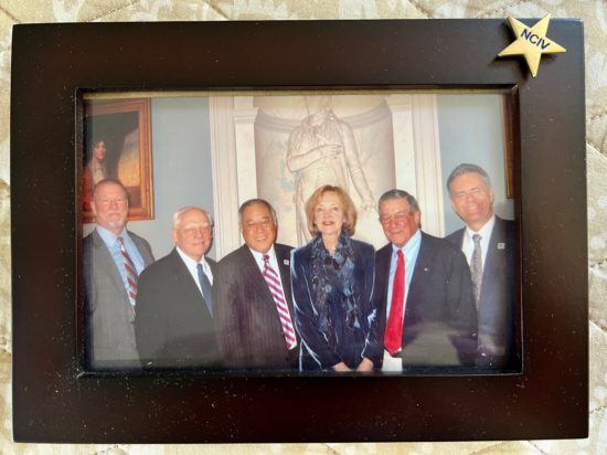 Group of people smiling at a photo in front of Roman artwork, and a picture frame along the edges of the photo with a sticker that says NCIV on it.