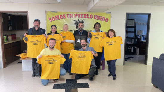 Group of people holding up yellow shirts in front of a sign reading "Somos Un Pueblo Unido."