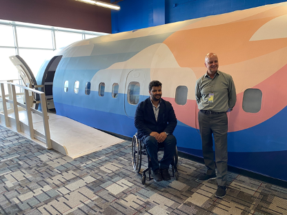 One man in wheelchair and one man standing pose in front of aircraft fuselage.