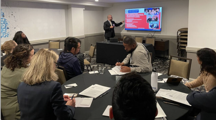 A male speaker stands in front of a powerpoint presentation he delivers to attendees seated at round tables in a hotel conference room.