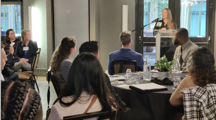 A blonde female speaker address attendees seated at round tables in a hotel conference room.